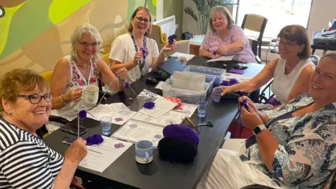 A group of women sit at a table making purple poppies.