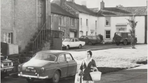 Hesket Newmarket Community Shop Black and white image of a woman in the 1960s walking through the village with shopping bags in front of the shop