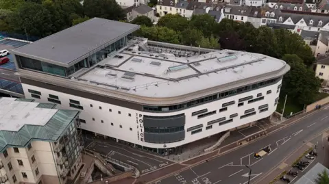 This is an aerial view of Jersey Police Headquarters, a large modern white building beside a main road, with surrounding residential properties, trees and a nearby car park visible from above.
