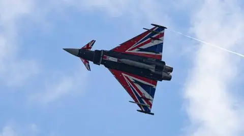 A typhoon flies in blue skies with white clouds, it has a pointed nose and triangular wings. It has red, white and blue paint, which reflects the union flag.
