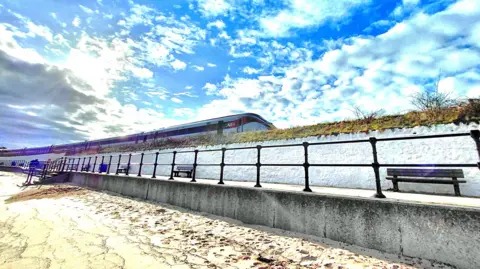 Stuart Miller The view from a beach on a sunny day, with a train speeding by just up from the beach.