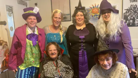 Six women wear an array of Halloween outfits, including a purple and black witch dress with pointy hats, a blue and white sequined dress, and a green plaid top and pink blazer 