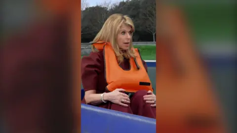 Kate Garroway is wearing a maroon top and bottoms, and an orange life jacket as she sits inside a blue pedalo 