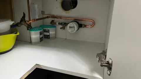 The inside of a kitchen cupboard in Swindon - stained from water damage - with a smart meter installed and de-humidifiers positioned next to it. 
