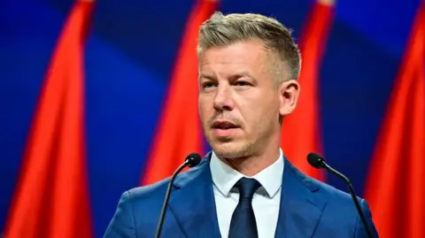 Magyar speaking into microphones with flags behind him. He is wearing a navy blue suit.