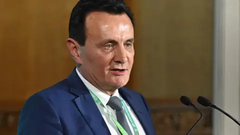 PA Media Pascal Soriot wears a blue suit, white shirt and grey-looking tie, standing in front of a microphone. He has very dark hair, slicked back, and is standing inside.