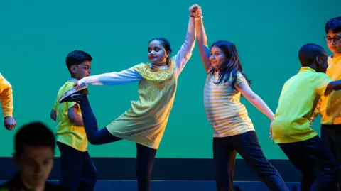 Elly Welford Two girls in yellow tops dancing with their arms up -holding hands, with boys dancing around them