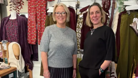 Emma standing with a second woman in the middle of rails of clothes in her shop, both are smiling to camera
