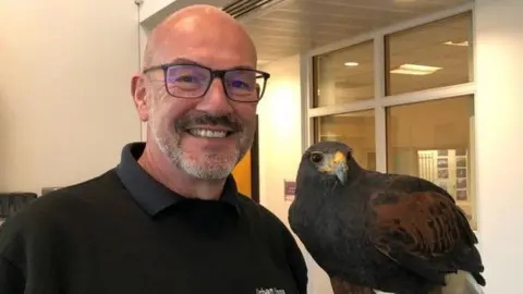 Andy Crowle smiling for the camera with one of his Harris hawks, Willow. They are inside a building.