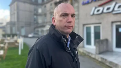 BBC Close up of Louis Gardner. Mr Gardner is wearing a black jacket and is looking to the right of the camera. He is standing outside. A large grey building is in the background.