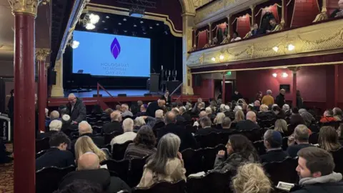 A theatre auditorium. People are sitting in rows, looking towards a stage. There is a projection on a screen saying, "Holocaust Memorial Day". The walls are maroon and gold.