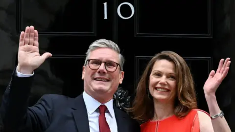 Getty Images Sir Keir Starmer, in a navy blazer, red tie and white shirt, and his wife Victoria, in a red dress, wave outside the door of 10  Downing Street