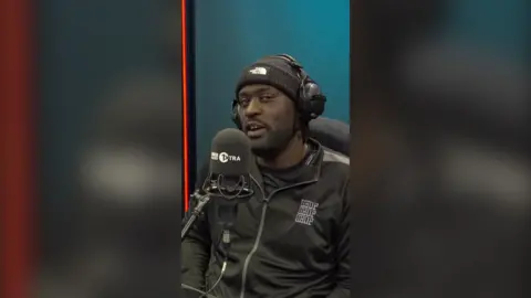 Rapper Blacks speaks into a microphone. He wears a black beanie hat and zip jacket