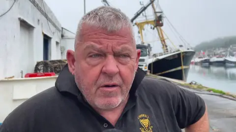 Fisherman Phil Mitchell wearing a black Cornwall branded polo shirt. He is stood on the harbour side and looking away from the camera. 