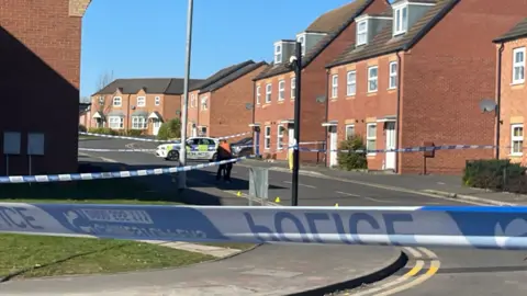 Police tape blocks off a street. A police car is parked in the road near some houses.