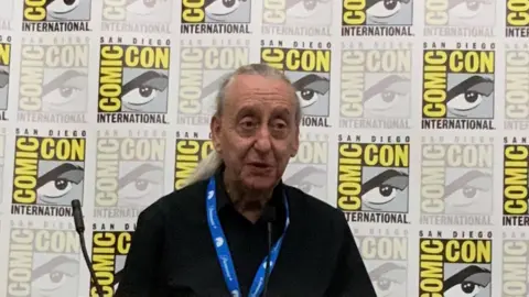 A man with long silver hair in a dark shirt stands on a stage with the San Diego Comic Con logo emblazoned across a panel behind