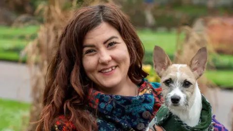 Samantha Morgan smiling at the camera, holding a small dog, she has dark long hair. In the background are plants.