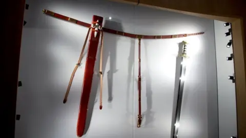 A display case showing a replica of the Sutton Hoo Sword, its scabbard and belt. The belt is at the  top of the display and hanging from it is the scabbard, with its two bosses mounted in white, and beneath the right end of the belt is a replica of the sword