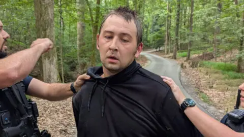 The Facebook/Jackson Police Department arrested Austin Robert Drummond, wearing a black hoodie, held by two policemen on roads in a wooded area. He doesn't look in front of the camera.