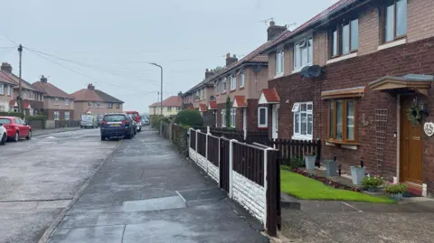 A street with semi-detached houses in what looks like a former council estate. The wide street has cars parked on the edge of the pavement on both sides. There are numerous two-storey houses with neat front gardens, hedges and fences.