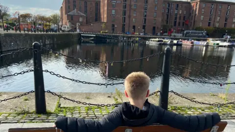 Boy with blonde hair sits on a bench looking at a dock with narrow boats in.