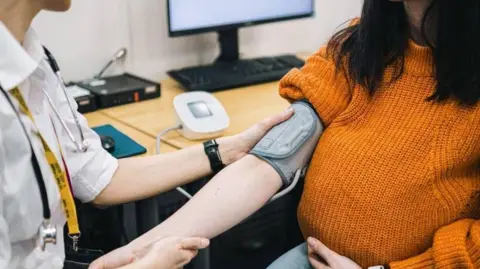 Getty Images A woman extending her arm out with a doctor measuring her heart rate with a device strapped to her elbow. She is wearing an orange jumper and has long hair down to her shoulders. A desk with a computer can be seen in the background.