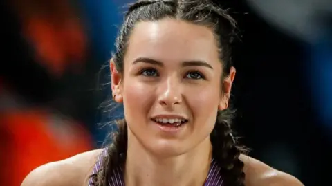 Getty Images A female athlete with a smile. She has dark hair in plaits and is in a a purple vest. She is wearing earrings. The background behind her is blurred but she appears to be in a sports hall or stadium.