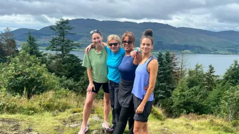 Duffy family Rachel Duffy wearing a green top, clack shorts and pink trainers. Amanda Collier is next to her wearing a blue top and black trousers. Judy Wilson and Roxy Wilson are both wearing blue sleeveless tops. They are standing in a wooded area with large trees. In the back ground you can see a large lough and a vast green mountain.