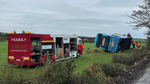 BBC A blue Ulsterbus lies on its side in a field. There are removal workers standing around wearing orange high visibility jackets and red removal truck is parked near the bus in the field. The damaged hedge can be seen in the foreground of the picture.
