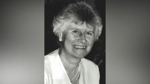 A black and white photograph of a smiling older woman, looking at the camera, with a pearl necklace and light blouse.