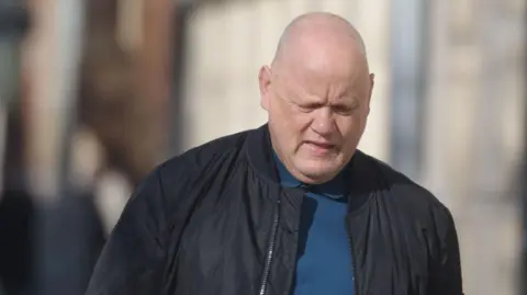 Mr Matthews is looking downward. He is bald and is wearing a black zip up cat and a blue t-shirt. 