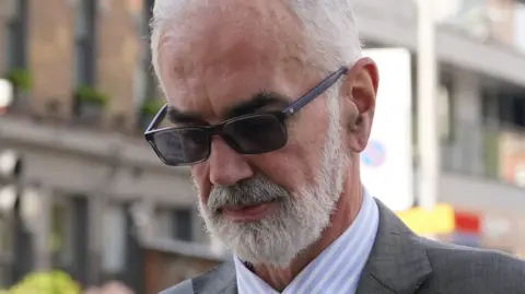A close up of the face of Cdr Julian Bennett, wearing sunglasses - he has a grey beard and grey hair and he is looking down. 