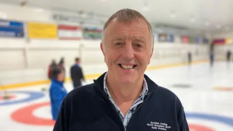 Keith Prentice, man in 70s with fair hair smiling, pictured with ice rink in background.