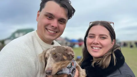 Richard Knights/BBC Kieran Smith, a man with dark hair and is wearing a cream coloured jumper. He is holding a brown and white dog which is wearing a grey harness. He is stood next to Megan Jones, a woman with mid length brown hair and blonde streaks who has a pair of sunglasses on her head and is wearing a black jacket.