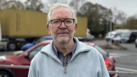A wide shot of Peter Dowd standing in front of a busy road. He has short white hair and is wearing a pair of glasses, a grey jumper, and a stripey shirt underneath.