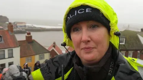 Michelle Lyons/BBC A woman who is a police officer standing on the steps in Whitby with a view of the harbour and some rooftops in the background. She wears a black police beanie hat and a yellow hi-vis jacket with a hood up. 