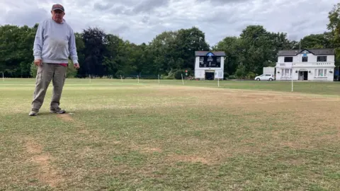 A man in a grey cap and light grey sweatshirt and trousers standing on a yellowing grass cricket pitch with faint bike skid marks leading up to his feet. There is a white cricket pavilion in the background.