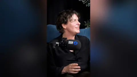 Woman with dark brown hair wearing a black shirt sat in front of a mic.