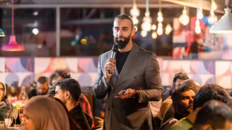 CN Photography A man in a grey pinstriped suit jacket and black jumper standing up and holding a microphone 