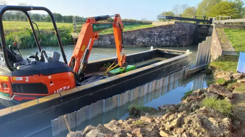 A barrier separates the towpath and canal debris from the main waterway where a machine sits on a low barge ready to dig. It is by a lock.