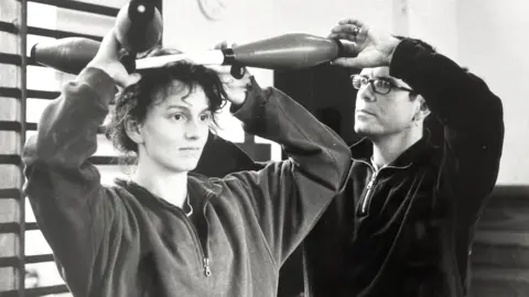 Circomedia A teacher guiding a student who is balancing juggling clubs on her head. It is an old black and white photo.