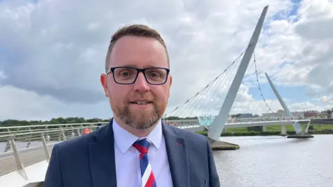 BBC Gary Middleton wearing navy suit, white shirt and red, white and blue tie, pictured in front of the Foyle Peace Bridge.