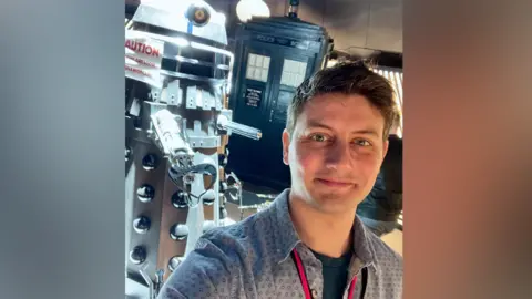 Scott Handcock Scott wearing a blue patterned shirt and red lanyard, standing in front of models of a Dalek and the Tardis and smiling at the camera