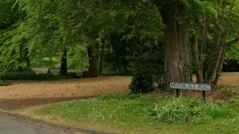 A street sign reading Melton Old Road, with a grassed and wooded area in the background and the edge of a road visible in the foreground.
