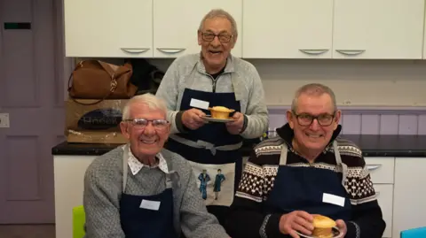 Three smiling older men in a kitchen wear aprons and hold two pies on plates