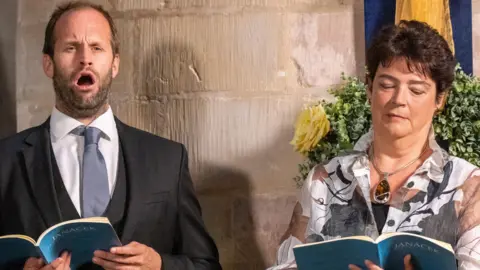 Dale Hodgetts A man and a woman performing during the Glagolitic mass at Gloucester Cathedral during the Three Choirs Festival. They are both stood up in front of a pillar and holding blue music books. The man has his mouth open, as he is singing. The woman is looking down at the book in front of her.