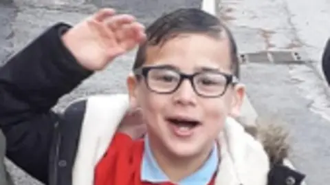 Family little boy with gelled down brown hair and black square framed glasses. He is smiling and waving 