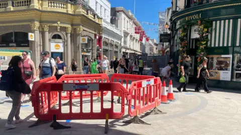 The King Street and New Street junction on a sunny day. There are plastic barrier fences that make a circle and a couple of traffic cones next to them. There are many pedestrians walking up and down the streets.