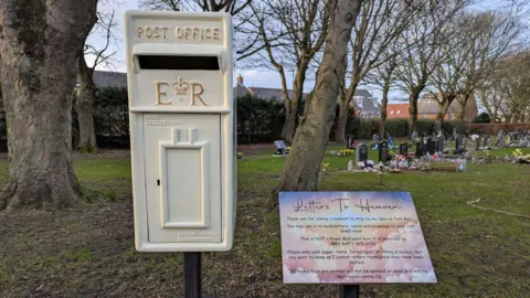 A white postbox sits on a pole in a cemetery. Next to it, a sign reads: "Letters to heaven".