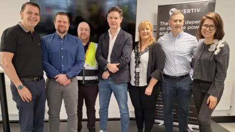 A number of people standing side-by-side in a line near a Vertiv advertisement